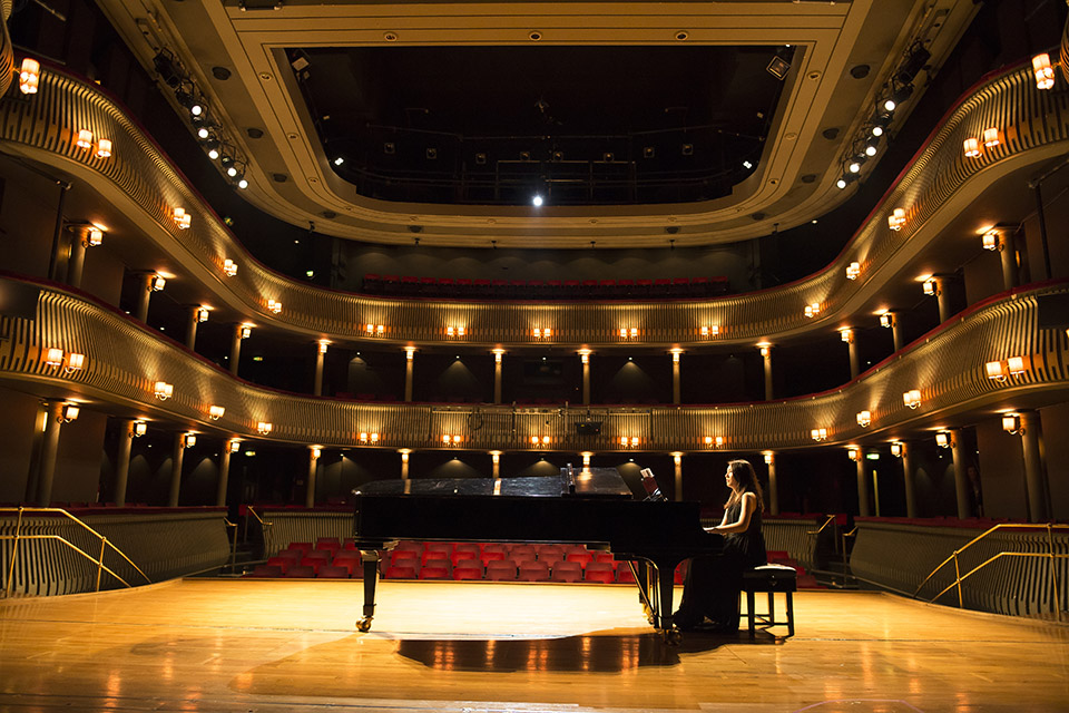 A female student, performing on the piano, on stage, in front of a brightly lit empty theatre.